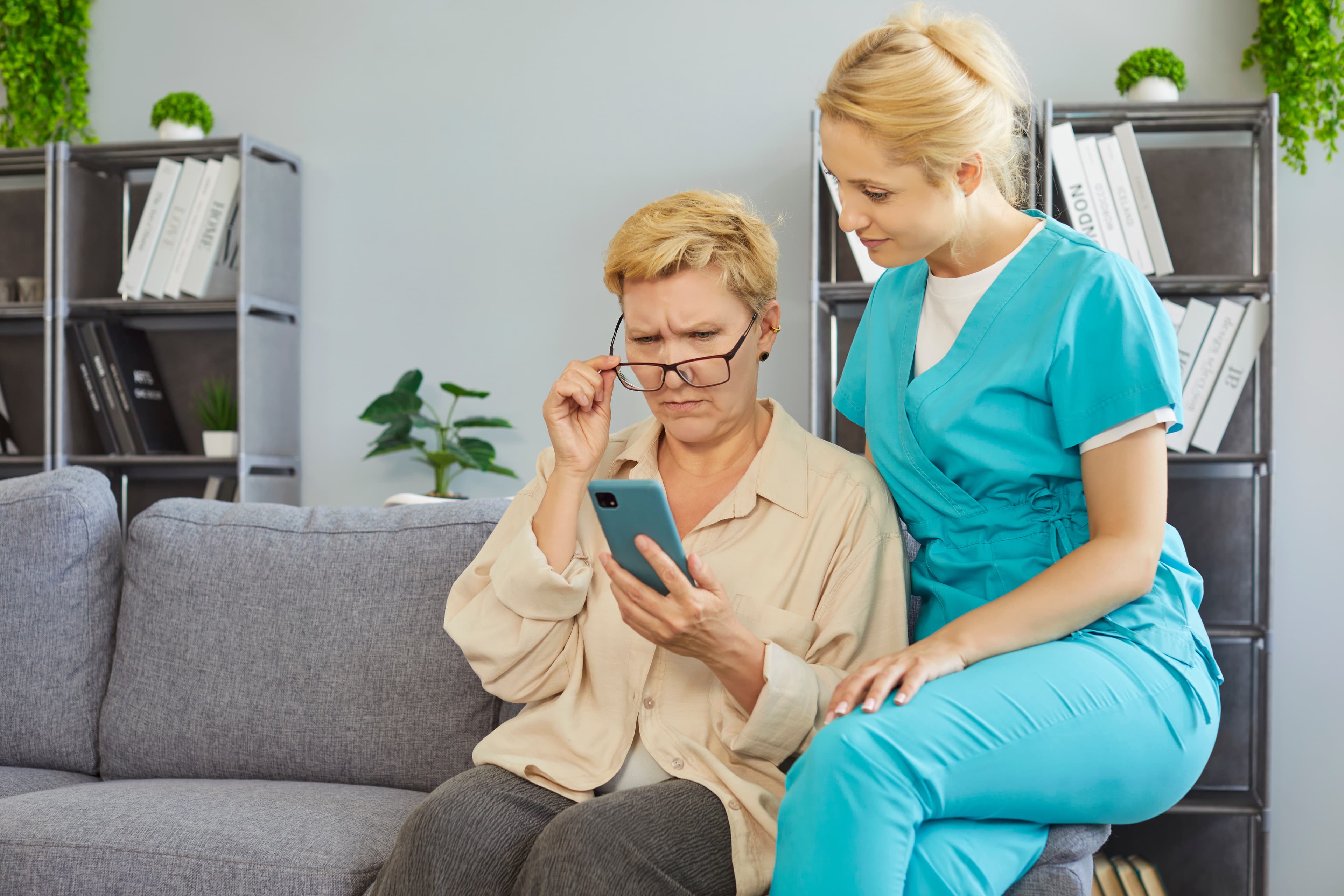 An older woman and a nurse in teal scrubs looking at a phone together on a couch