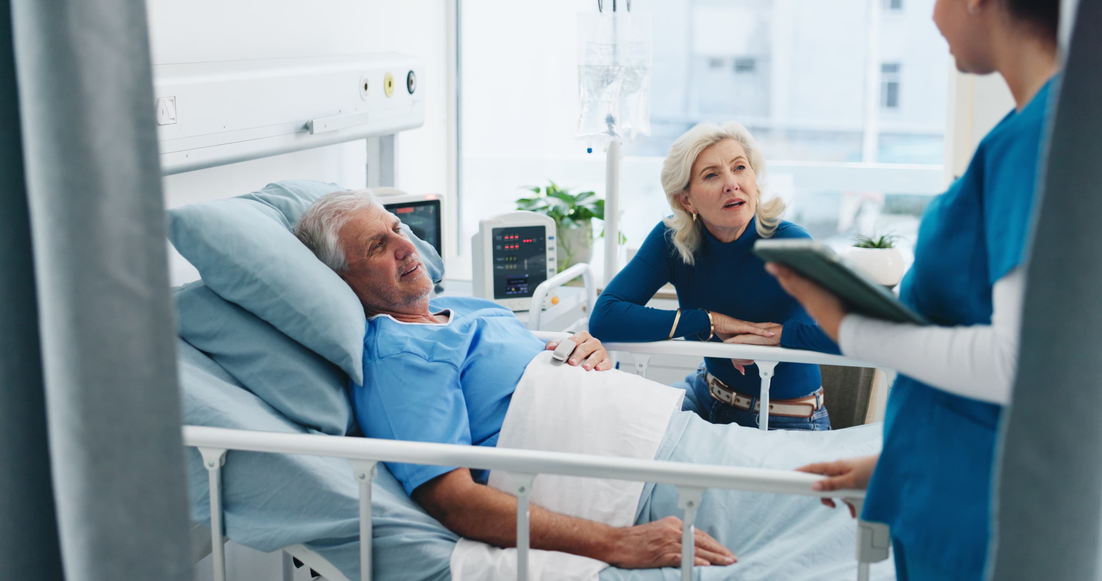 An older man in a hospital bed with his wife beside him while a clinician reviews notes on a clipboard