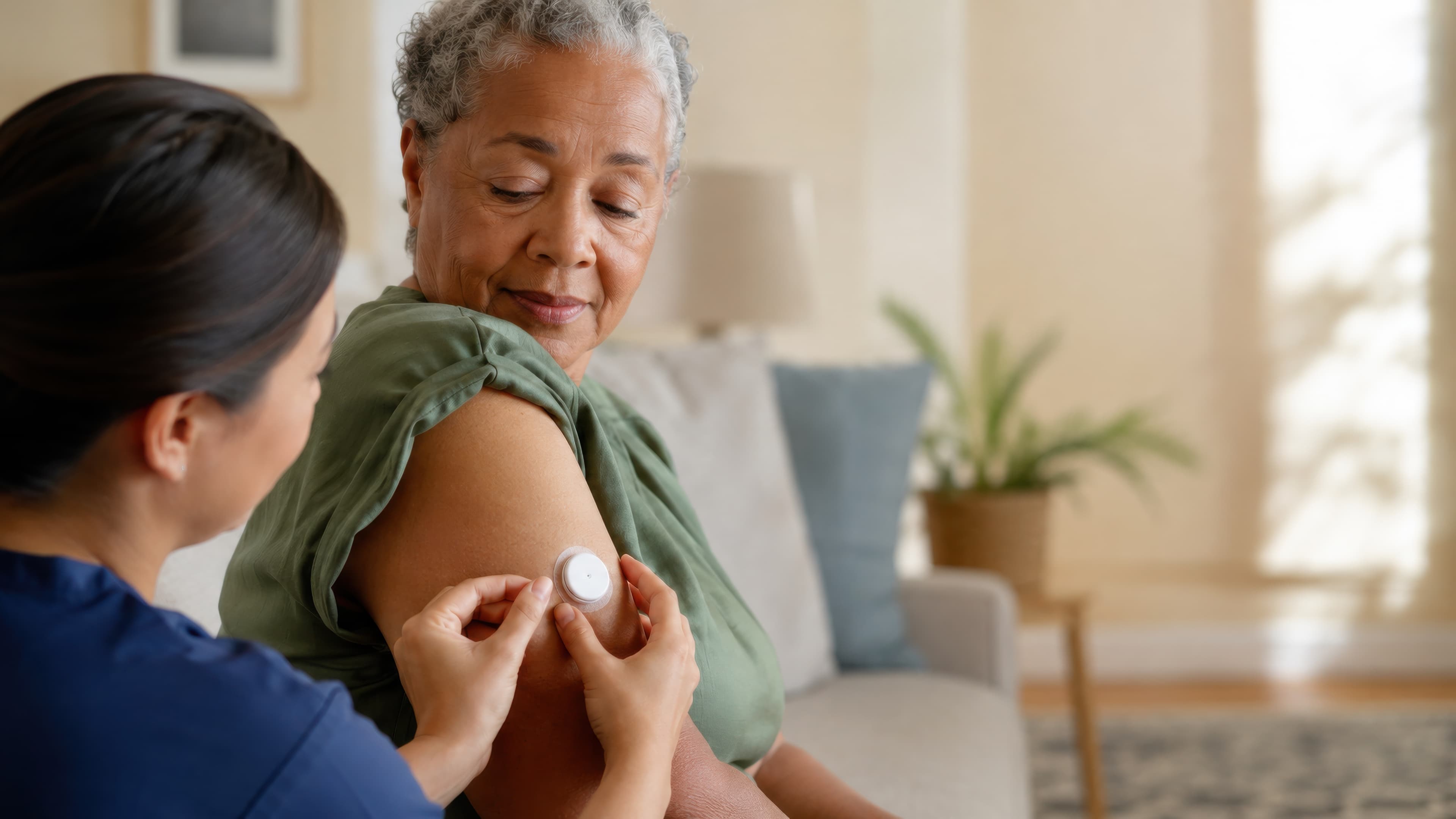 A nurse in blue scrubs placing a small round sensor on the upper arm of an older woman sitting on a sofa at home
