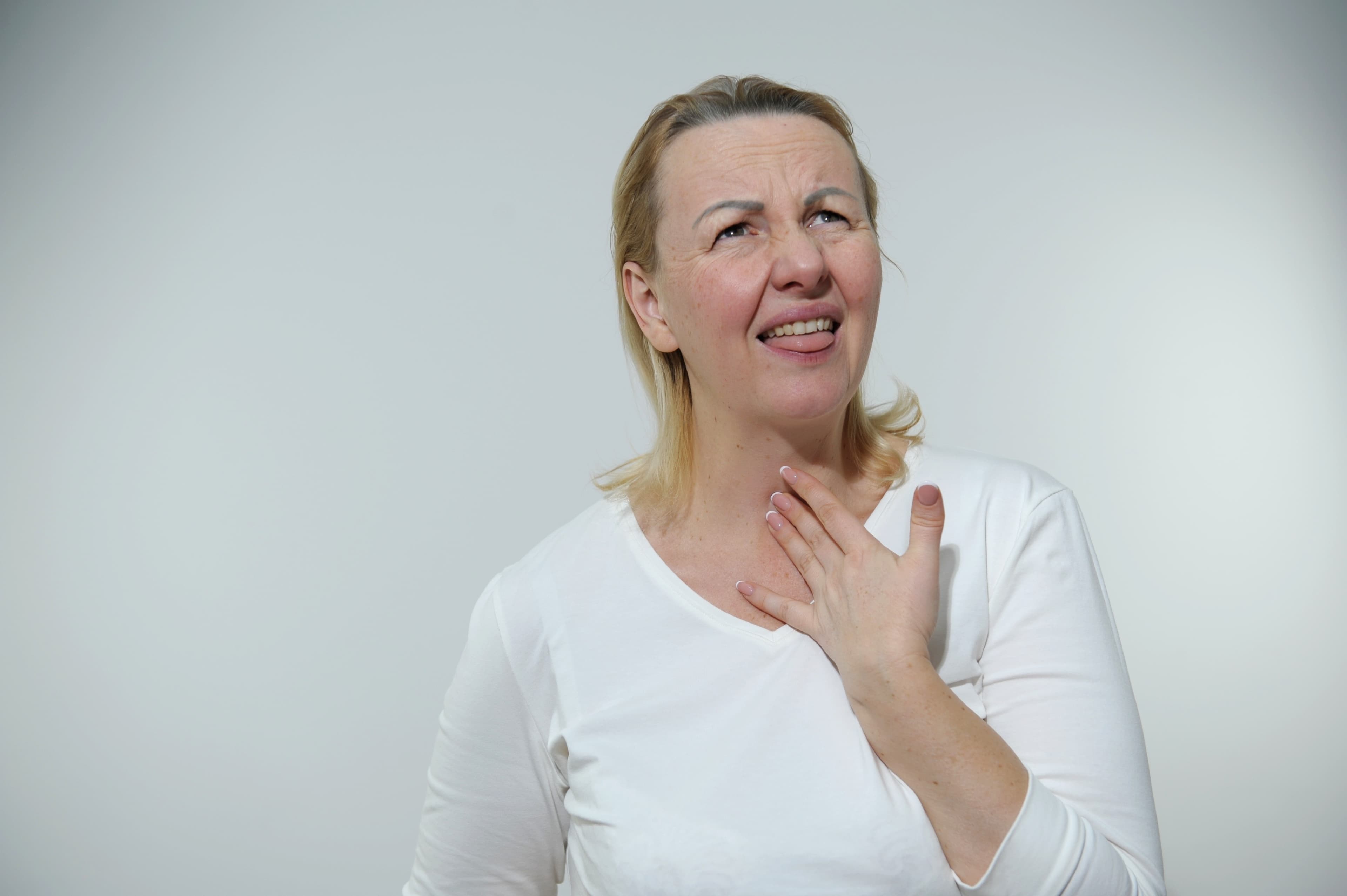 A woman in a white top with one hand at the base of the throat and a strained expression, against a plain gray wall