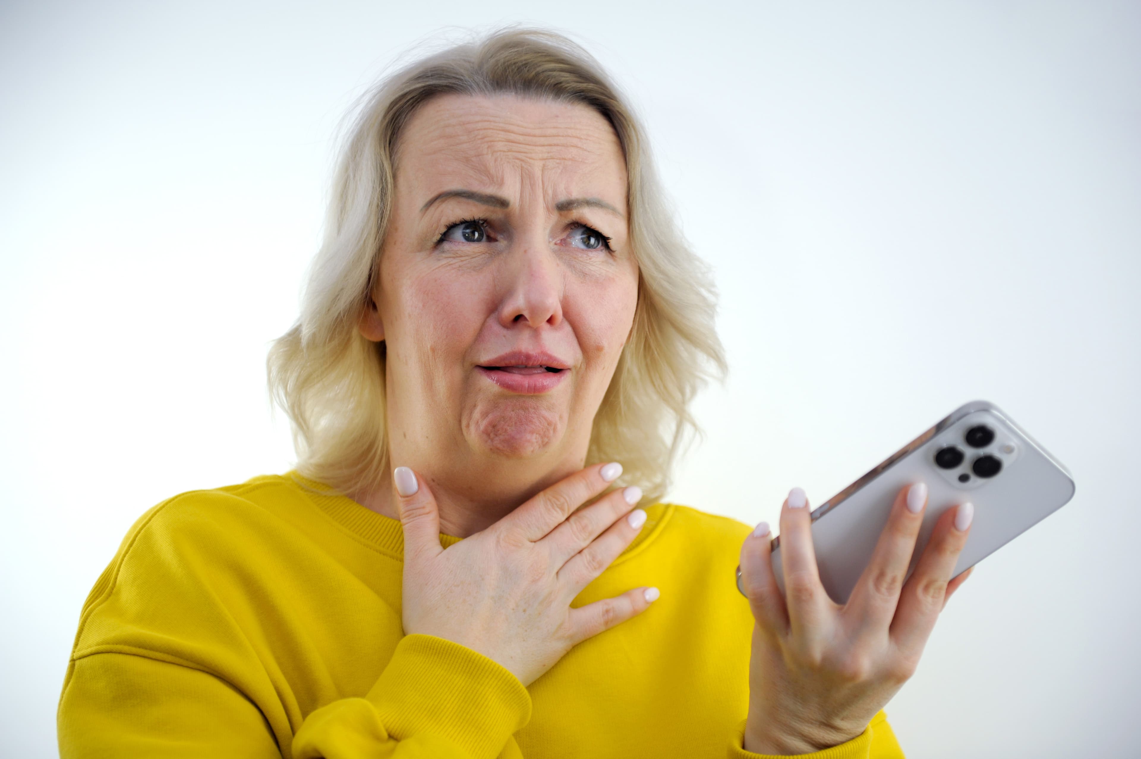 A woman in a yellow sweater holding a phone, her other hand resting on her throat