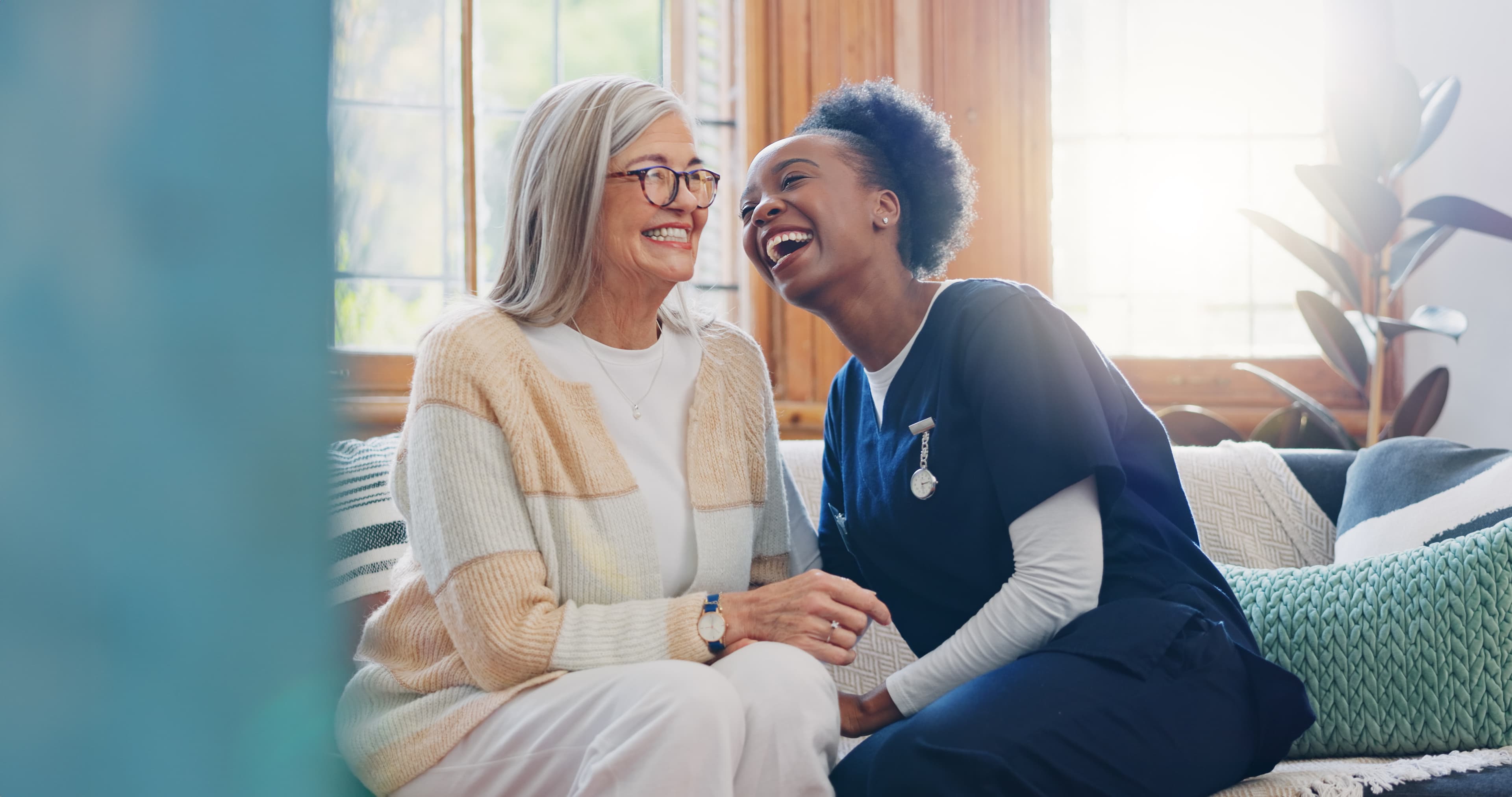 A home caregiver in scrubs laughing together with a smiling older woman on her couch
